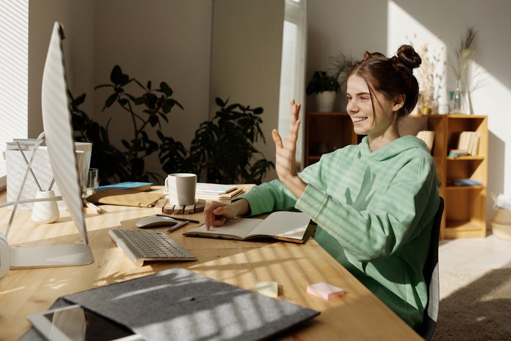 Cheerful woman in green sweater video calling from her home office, using a desktop computer.